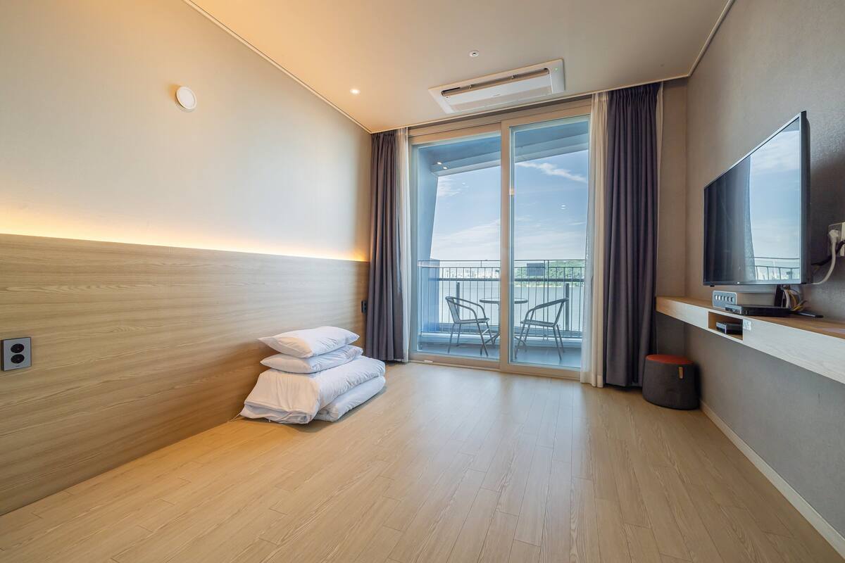 A modern hotel room with a large bed, a flat-screen TV, a small table, and a balcony with outdoor furniture.