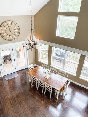 A dining area with a wooden table, white chairs, a chandelier, and a large wall clock. - Spring Lily Retreat - Spacious, Lakefront, Hot Tub, Private Dock, Peaceful Cove! (Speedwell)
