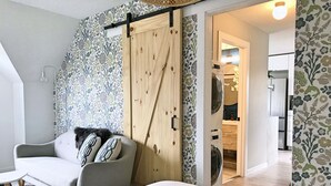 Ein modernes Schlafzimmer mit einer grauen Couch, einem flauschigen Teppich und einer Holztür, die sich als Schiebetür zu einem Badezimmer mit Waschmaschine öffnet.