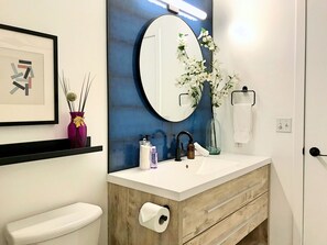 A bathroom with a white toilet, a wooden vanity with a mirror, a framed picture, and a vase with flowers. - Mountain View Escape - Top Floor (Canmore)