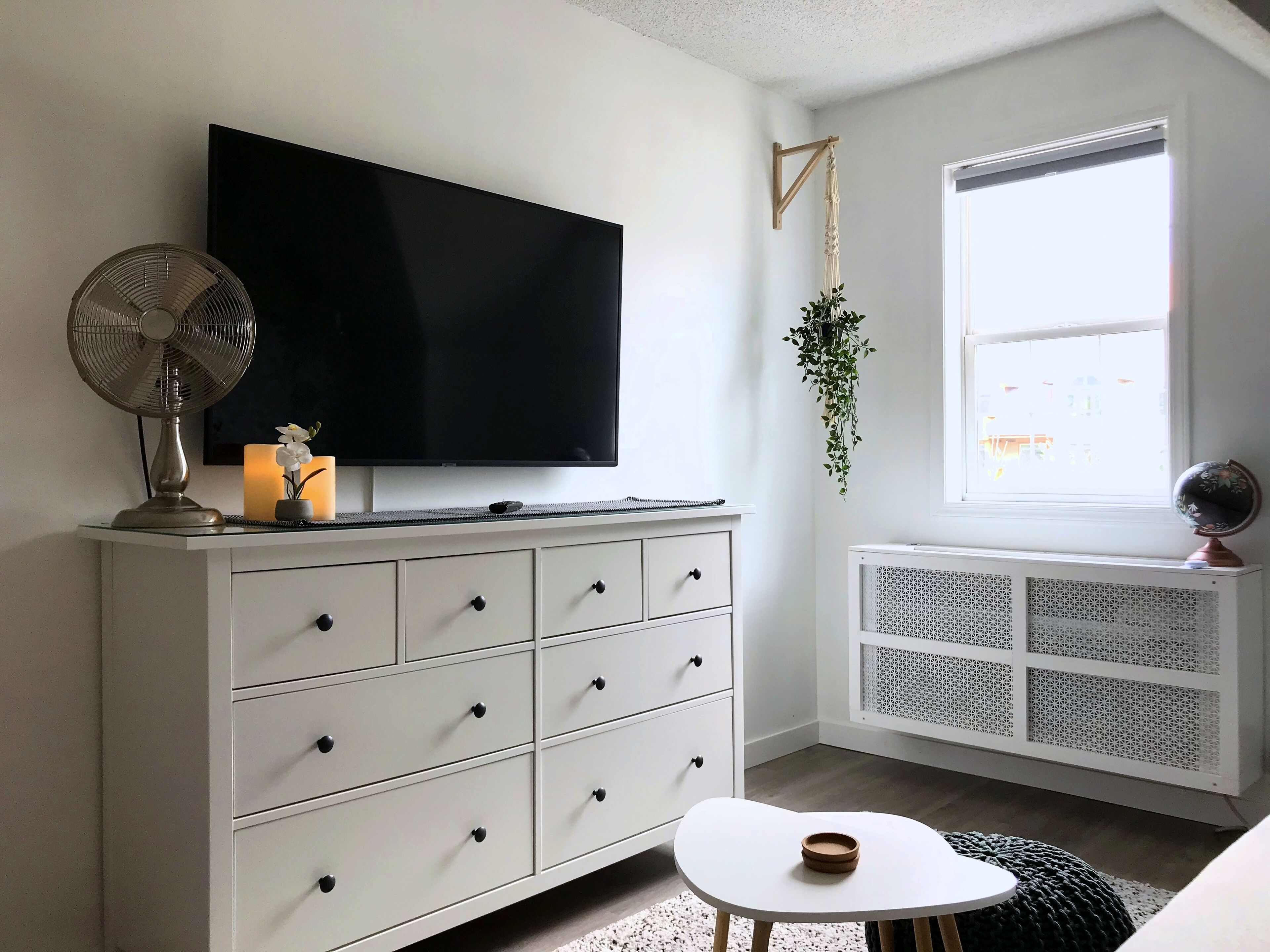 Una habitación moderna con un tocador blanco, un televisor de pantalla plana, un ventilador de pie, una mesita con un bol, una planta colgante y un radiador.