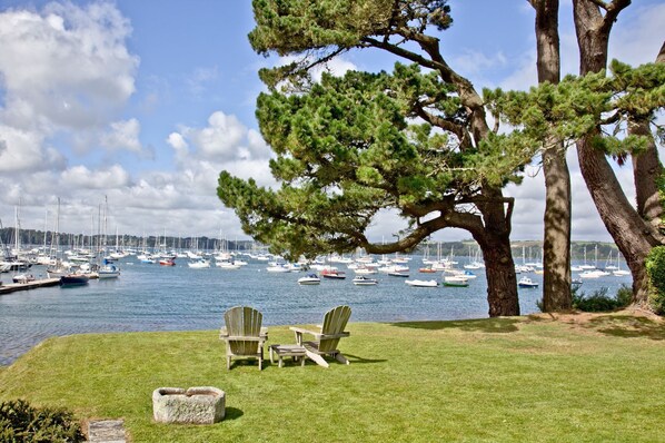 Een gazon met twee Adirondack stoelen en een stenen plantenbak, met uitzicht op een jachthaven vol zeilboten.