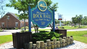 A welcome sign for Rock Hall with a boat graphic, a temperature display, and a bank sign in the background.
