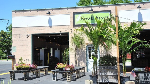 A building with a sign reading "Fresh Start" and a storefront with a sign that says "Fresh Food." There are outdoor tables with potted plants and an American flag.