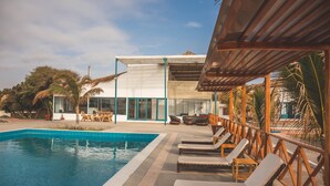Un área de piscina con sillones reclinables, un edificio moderno con amplias ventanas y palmeras.