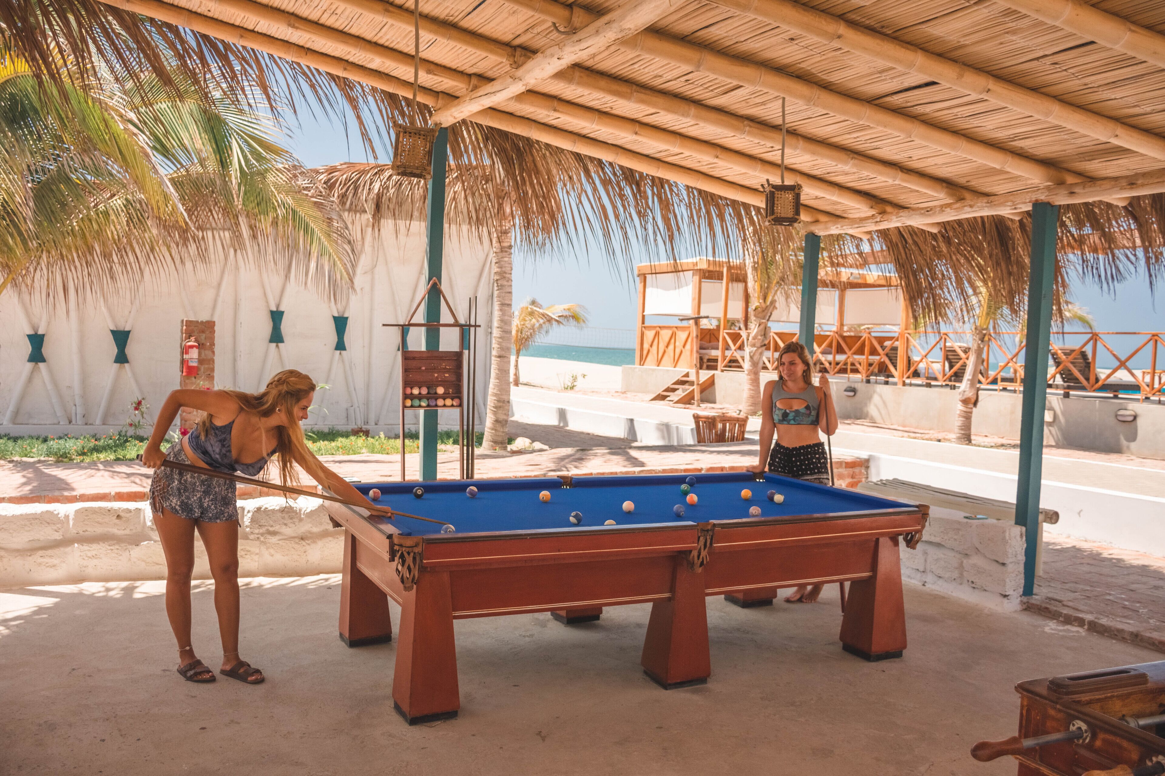 A pool table under a thatched roof with a view of the beach and ocean.