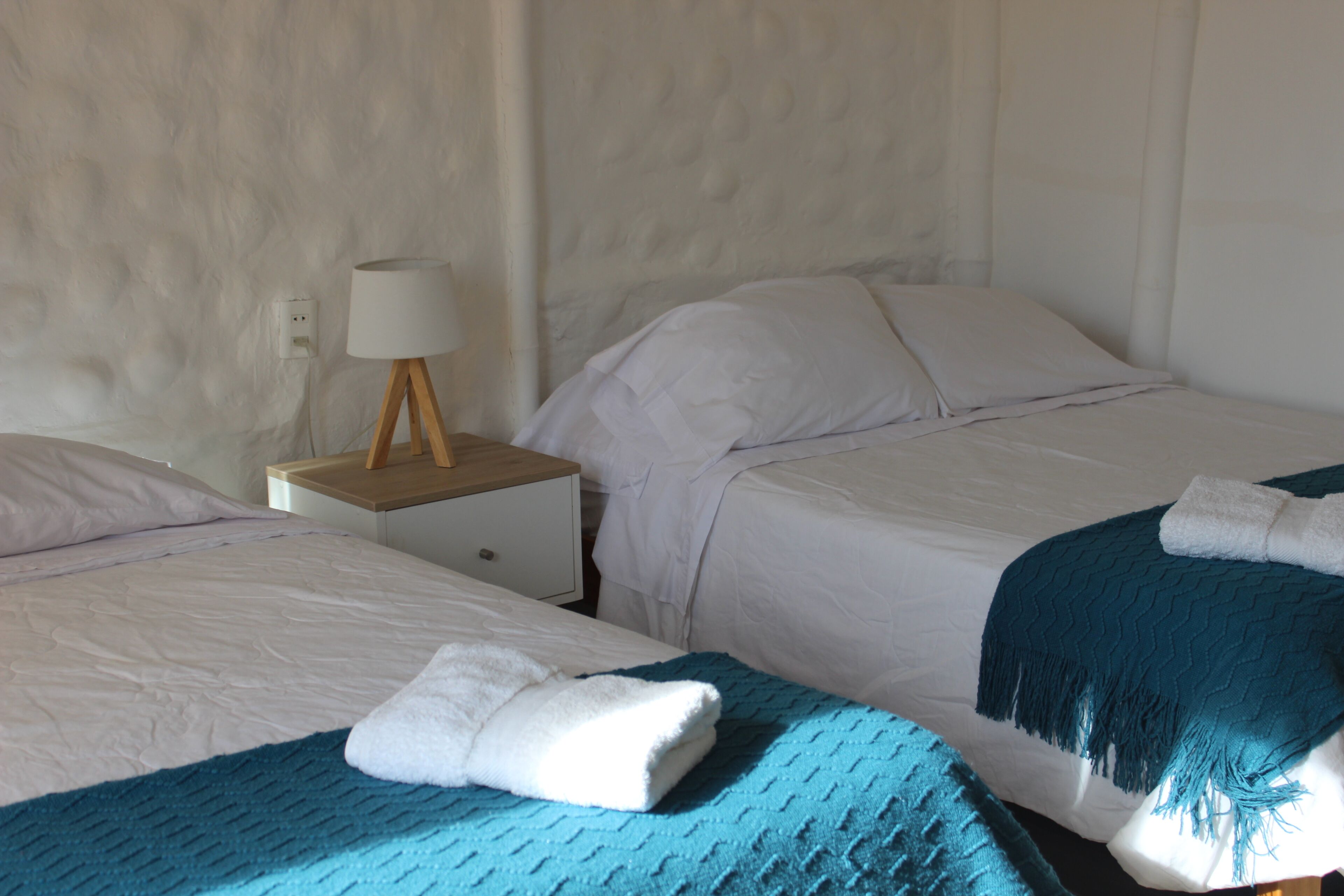 A hotel room with two beds, white bedding, a wooden nightstand with a lamp, and a blue throw blanket.