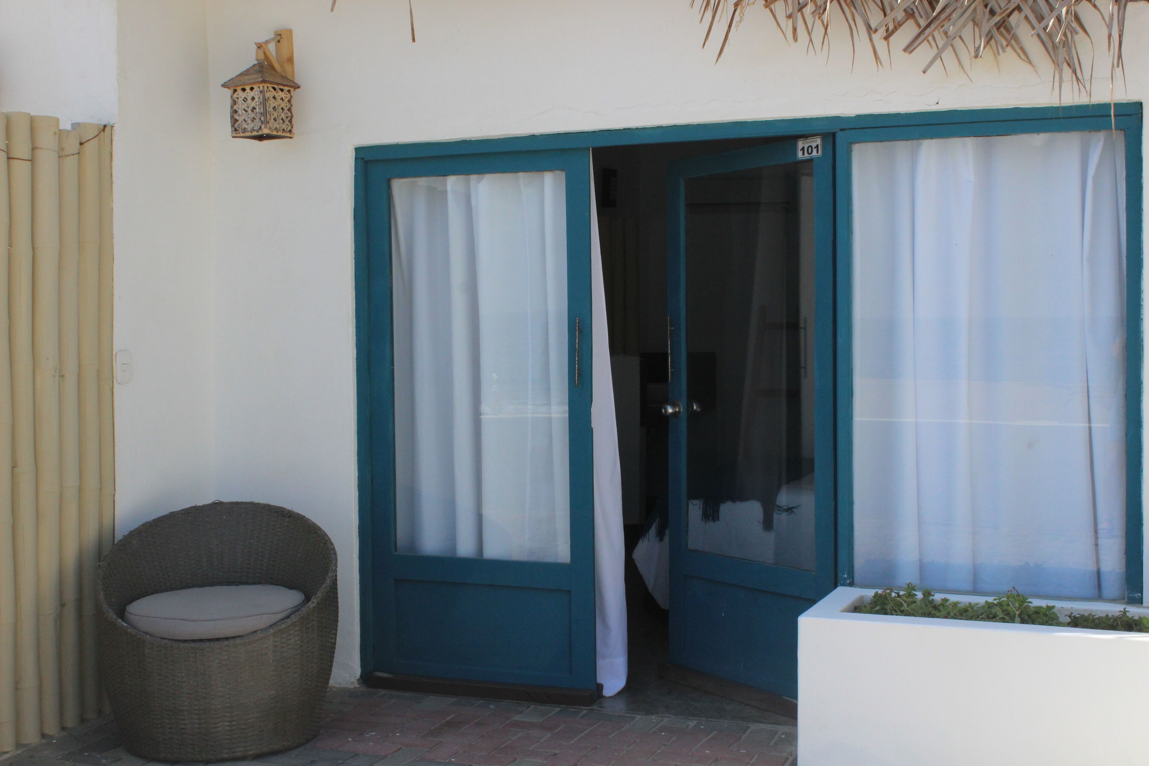 A room with a wicker chair, a white planter with greenery, and a blue door with a number sign.