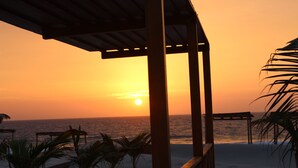 Escena de playa con una pérgola de madera, palmeras y vista al océano al atardecer.