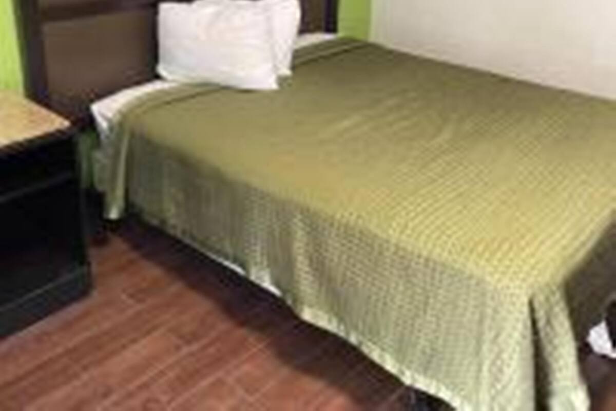 A single bed with a green bedspread in a room with a wooden floor and a white wall.