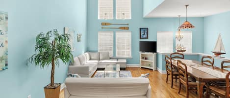 A living room with a sofa, coffee table, dining table with chairs, a potted plant, and a TV.