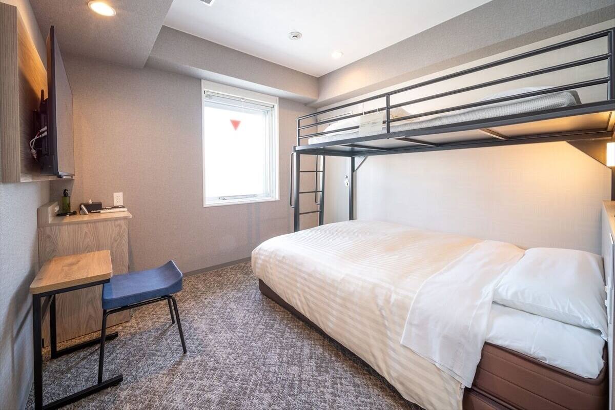 Pieni hotellihuone, jossa on kerrossänky, työpöytä tuolilla, pieni ikkuna ja patteri.