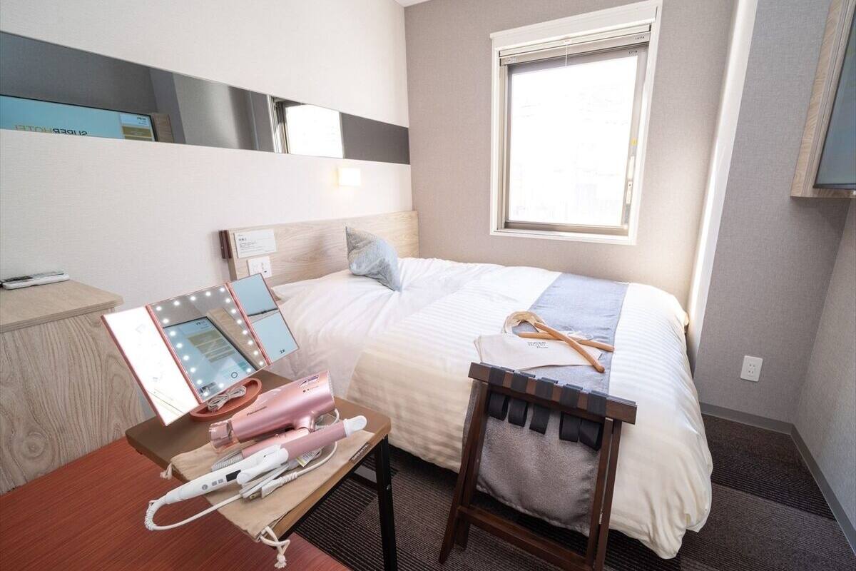 Hotellihuone, jossa on sänky, peilillä ja valoilla varustettu meikkipöytä, työpöytä kannettavalla tietokoneella ja ikkuna.