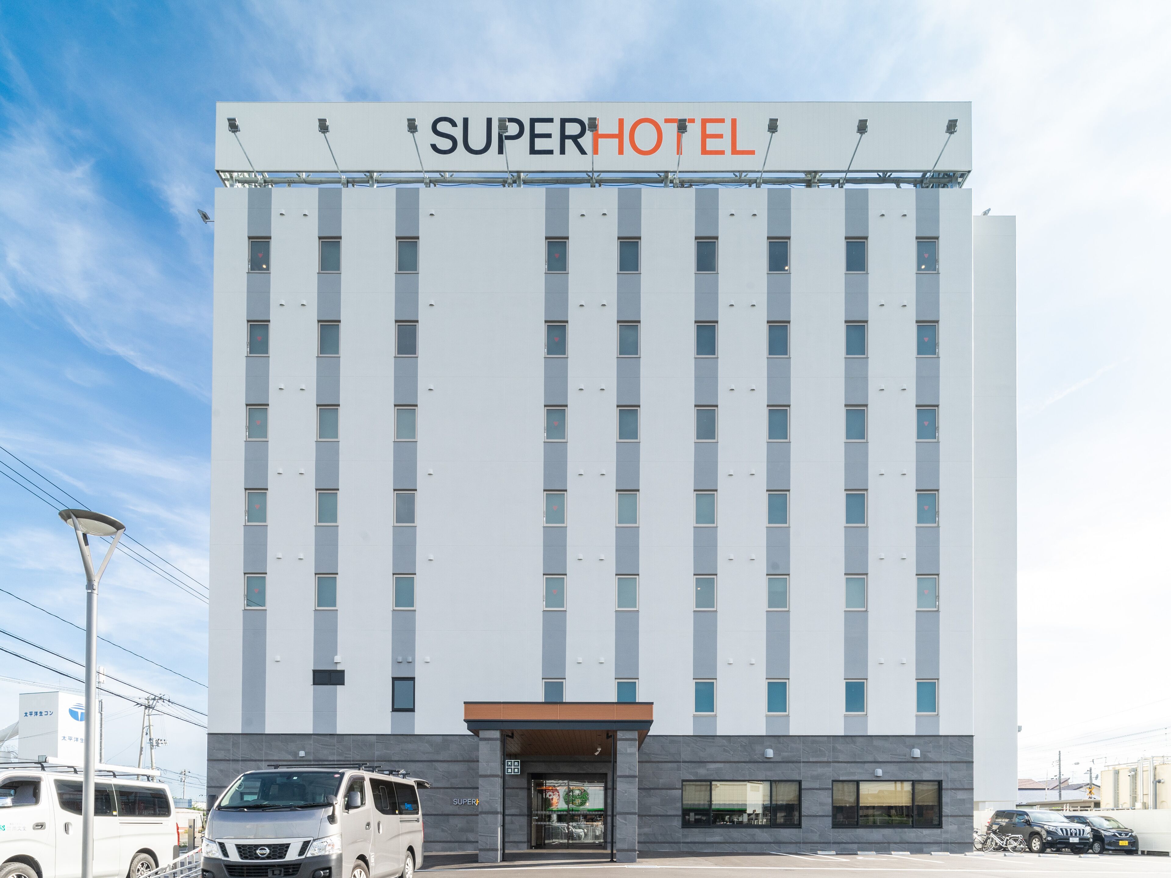 Un moderno edificio hotel con un cartello che riporta “SUPERHOTEL”. Presenta una facciata simmetrica con finestre regolarmente distanziate e un ingresso centrale.