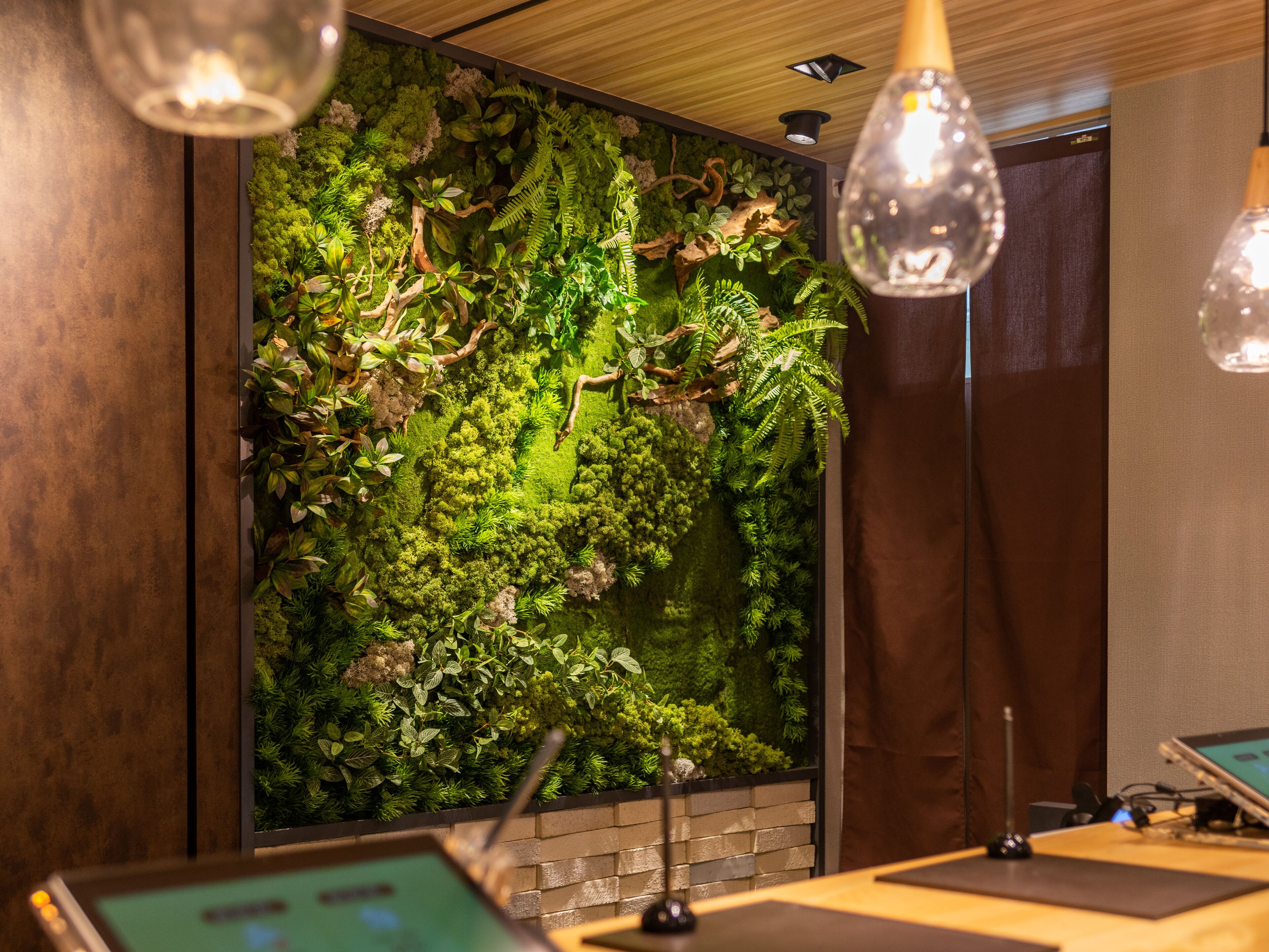 Una habitación con un gran muro vertical cubierto de plantas, un escritorio de madera con una computadora portátil y lámparas colgantes.