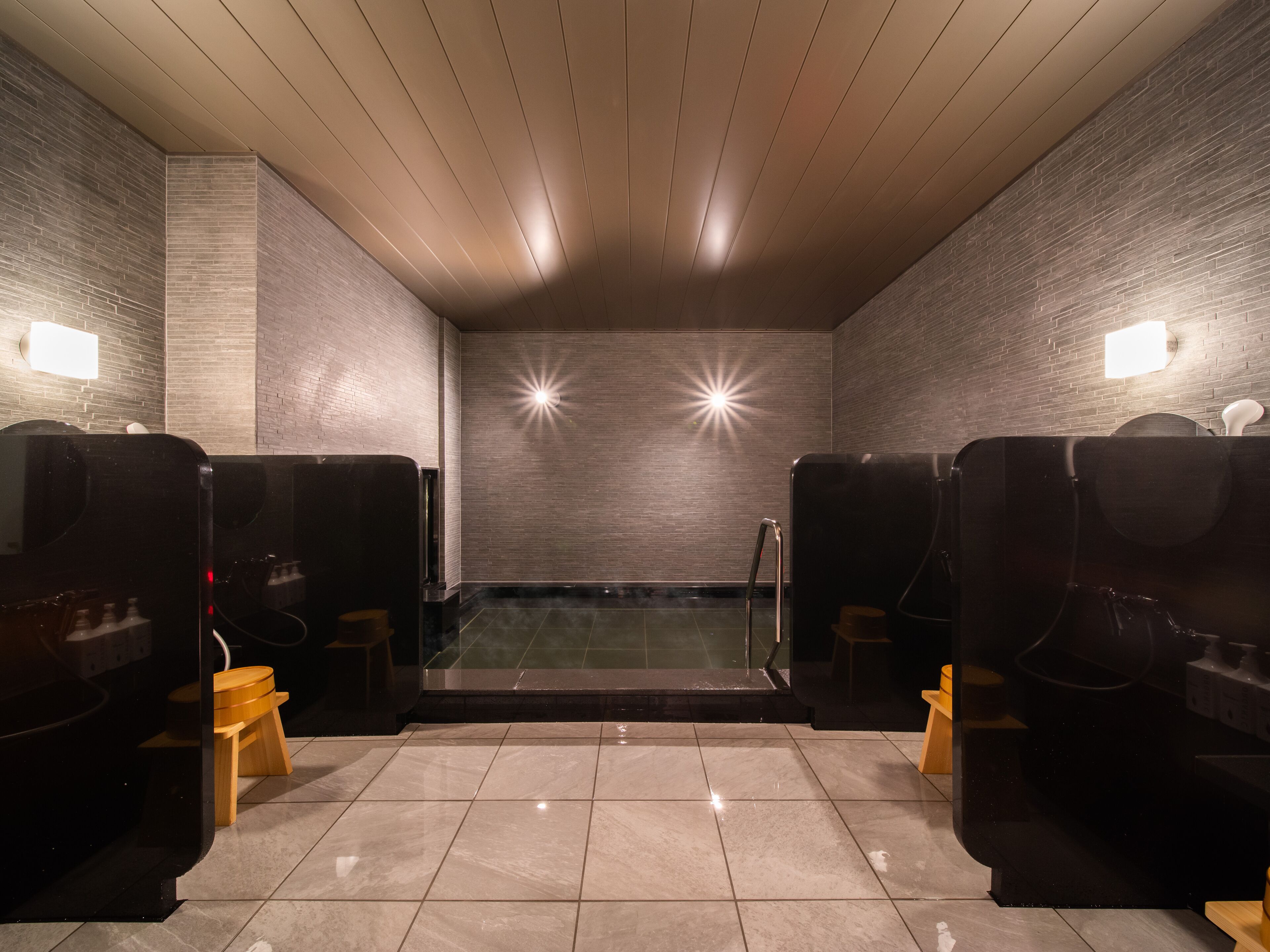 A sauna room with two wooden benches, a tiled floor, and a brick wall with two wall-mounted lights.