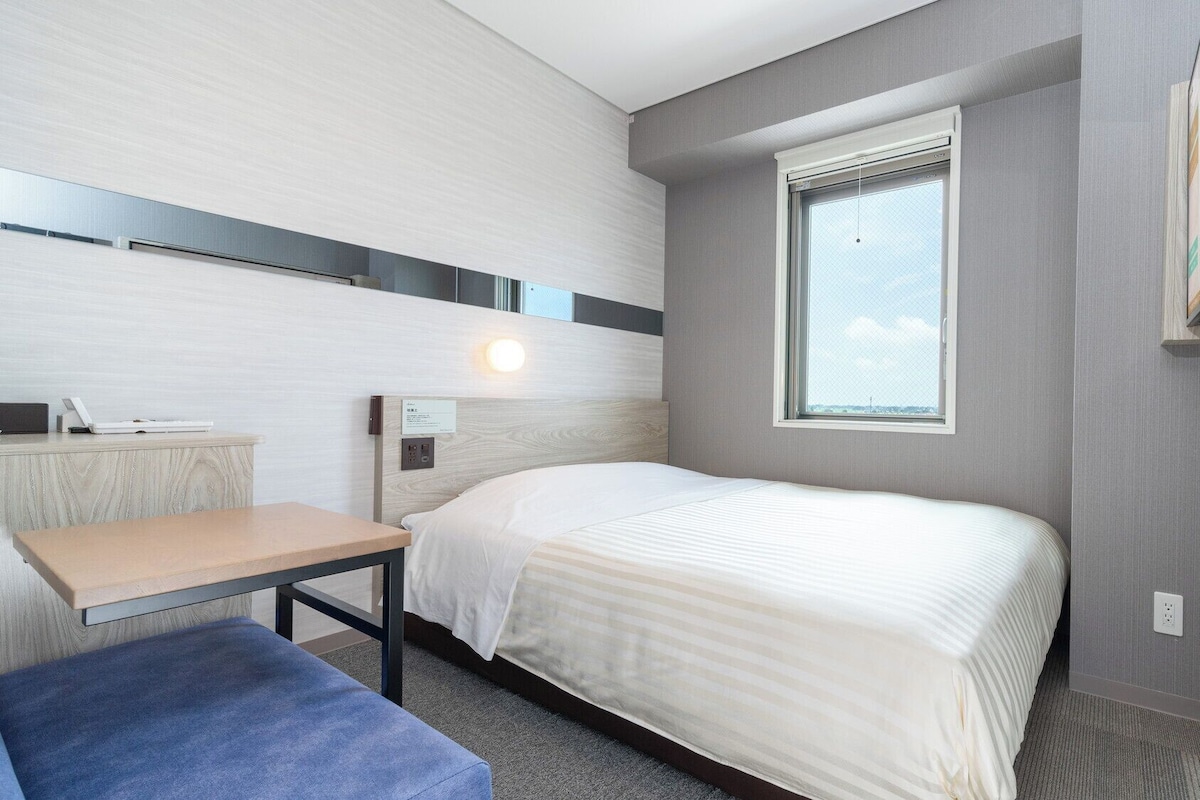 A compact hotel room with a bed, a desk, a chair, a window, and a wall-mounted TV.