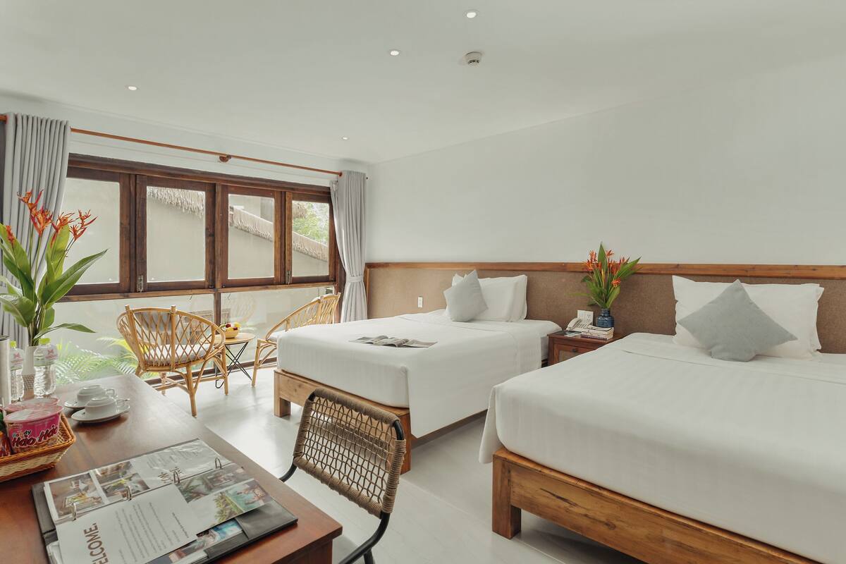 A hotel room with two beds, a dining table, chairs, a small table with a plant, and a window with curtains.
