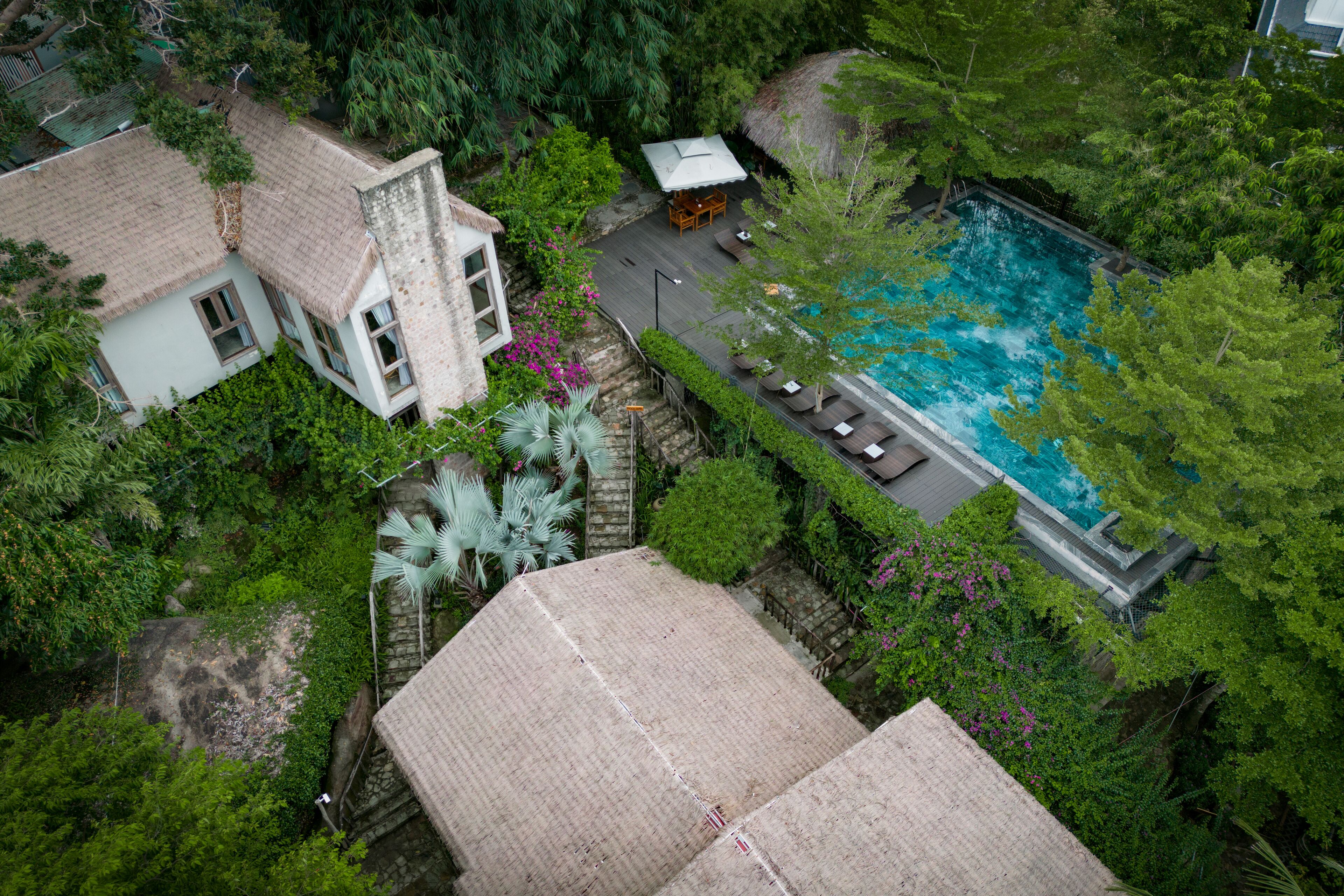 An aerial view of a property with a swimming pool surrounded by lush greenery, a thatched-roof building, and a paved area with seating.