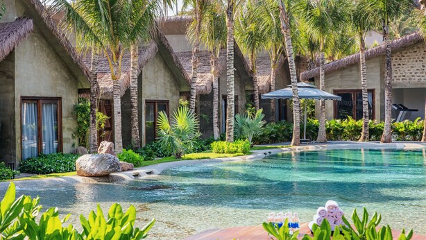 A pool area with a thatched-roof building, palm trees, and a lounge chair. - M Village Phu Quoc (Phu Quoc)