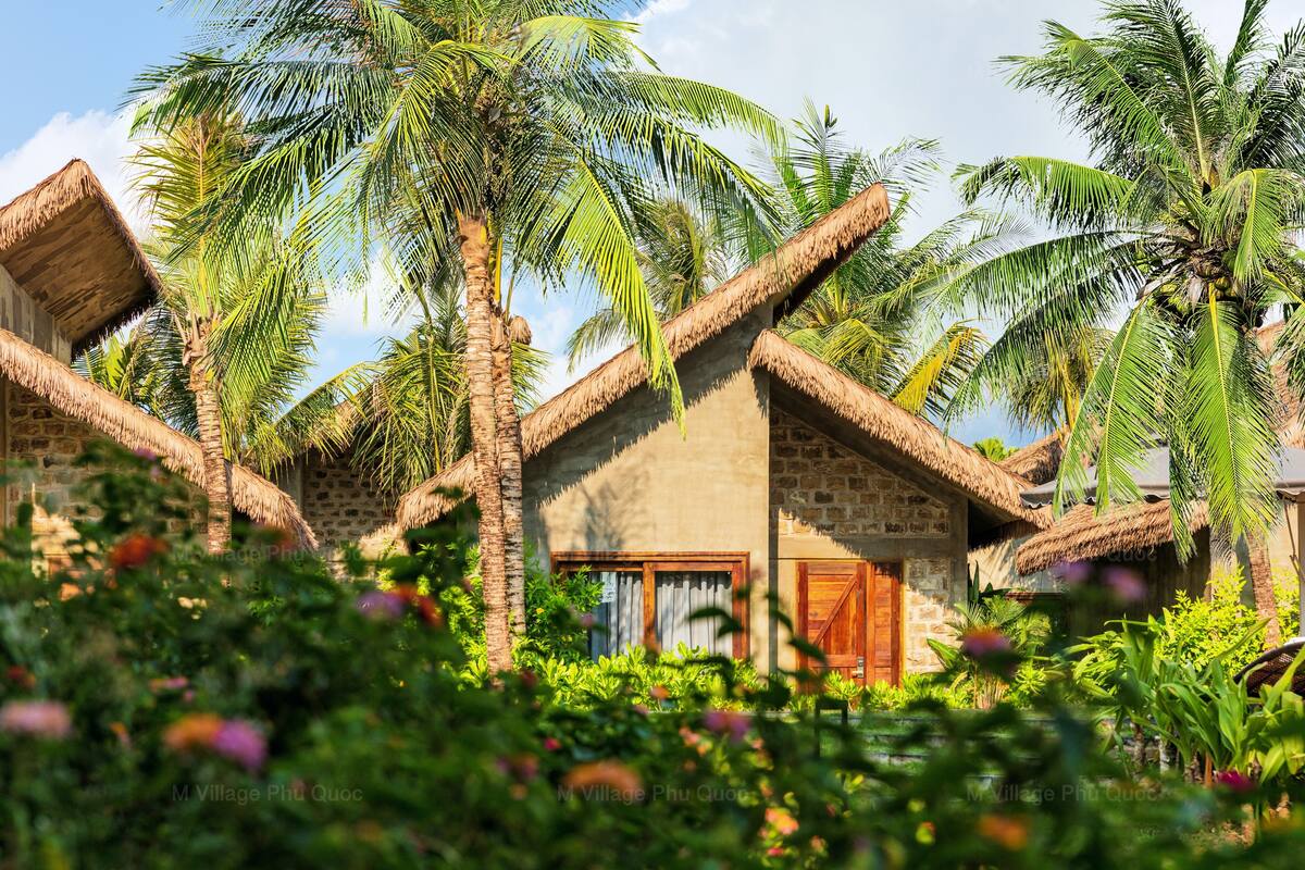 A traditional thatched-roof house with a wooden door and windows, surrounded by lush greenery and palm trees.
