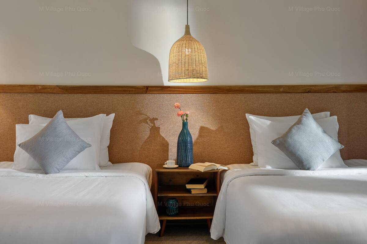 A hotel room with two beds, a nightstand with books and a vase, and a hanging light fixture.