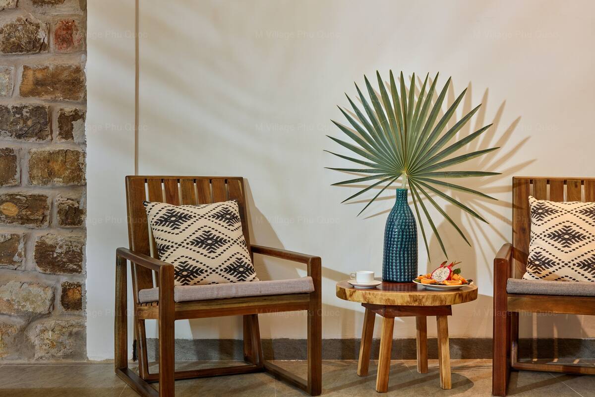 Two wooden chairs with patterned cushions, a small wooden table with a blue vase and a potted plant, and a brick wall.