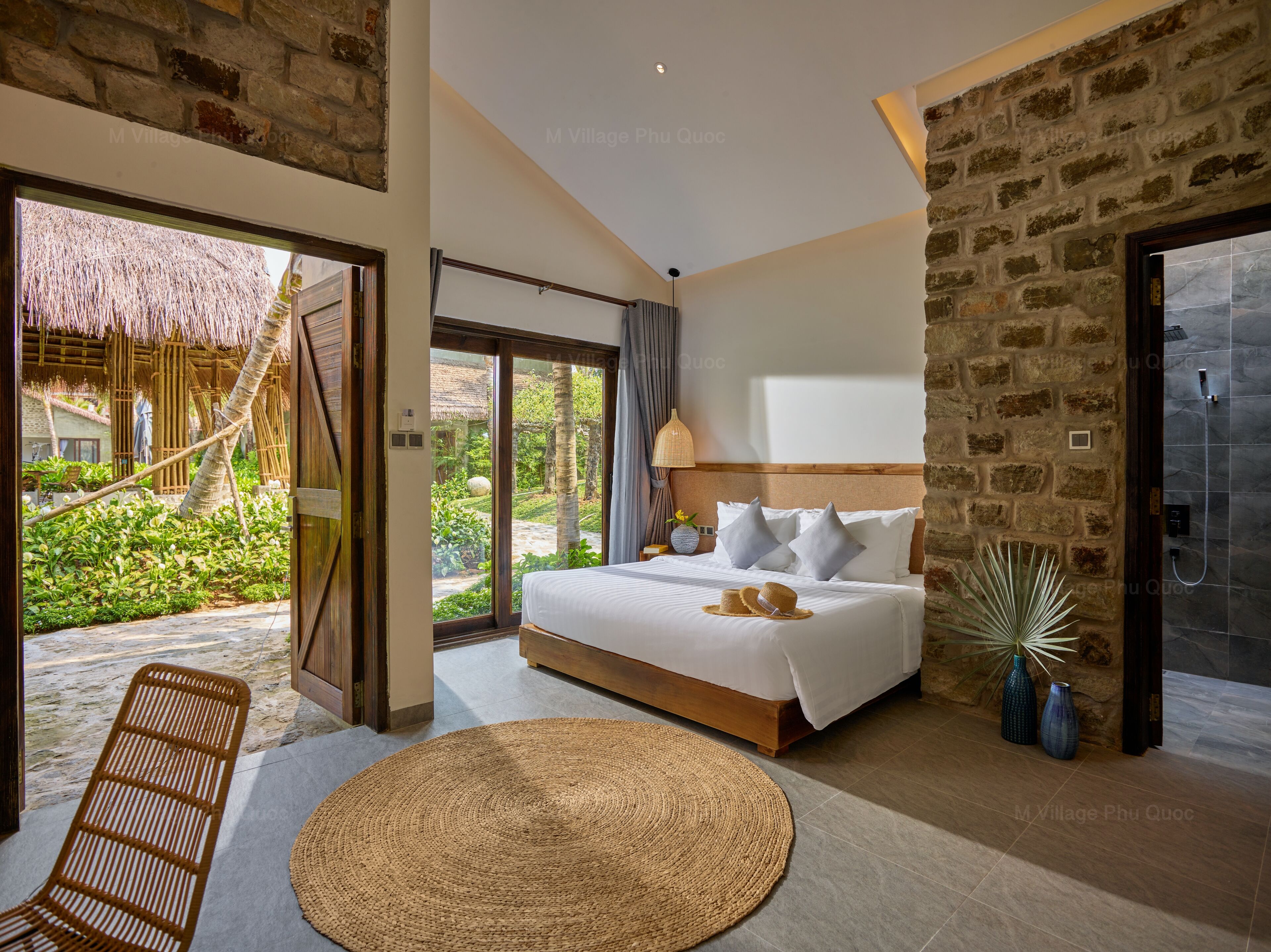 Une chambre avec un grand lit, une vue sur un jardin, un mur en pierre et une porte vitrée donnant sur l’extérieur.