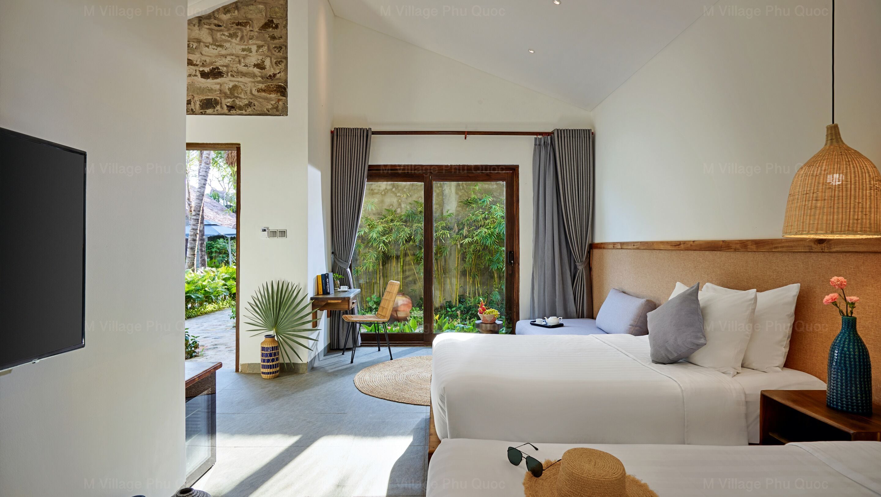 Una habitación de hotel moderna con una cama grande, otra cama, un televisor, un área de comedor con una mesa y sillas, y vista a un jardín a través de la puerta de cristal.