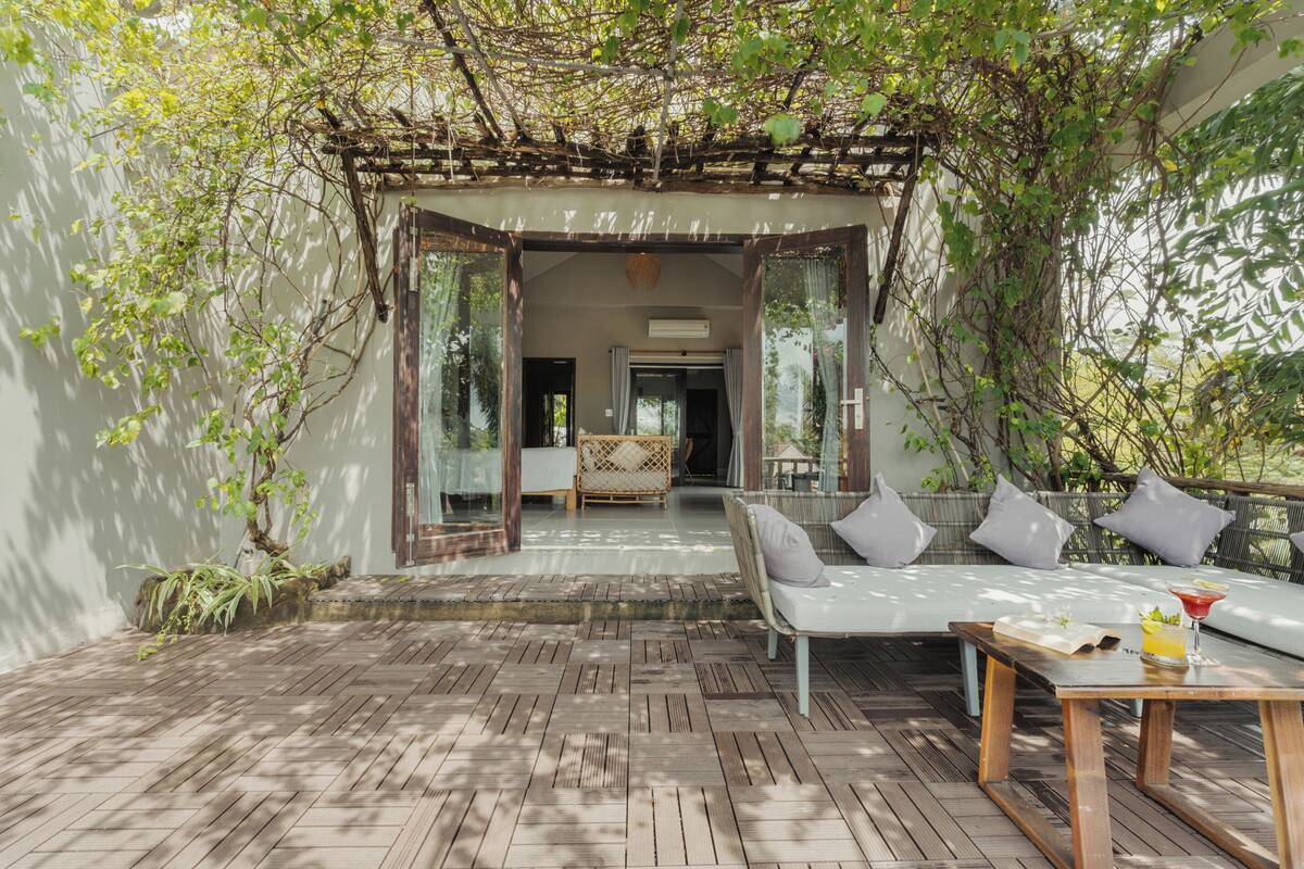 A covered outdoor seating area with a wooden deck, a sofa with cushions, and a table with a drink.