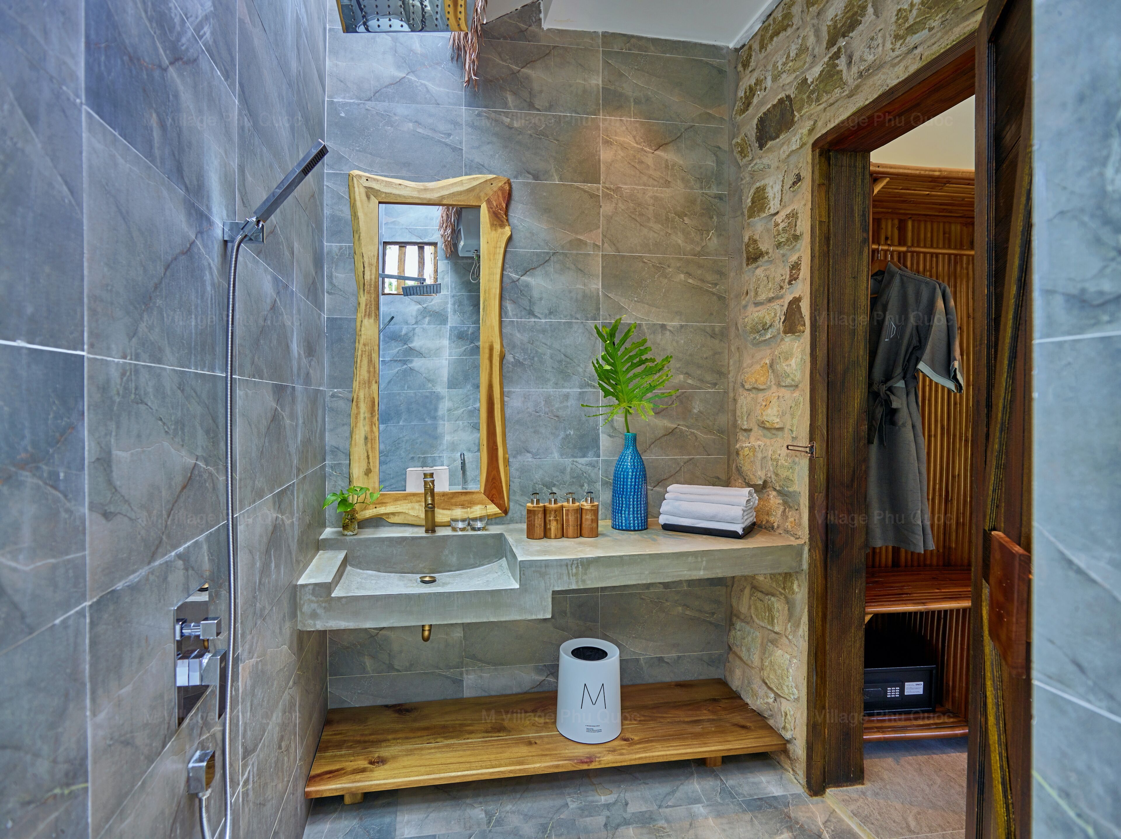 A bathroom with a stone wall, a wooden vanity, a mirror, a sink, a towel rack, a plant, and a shower area.