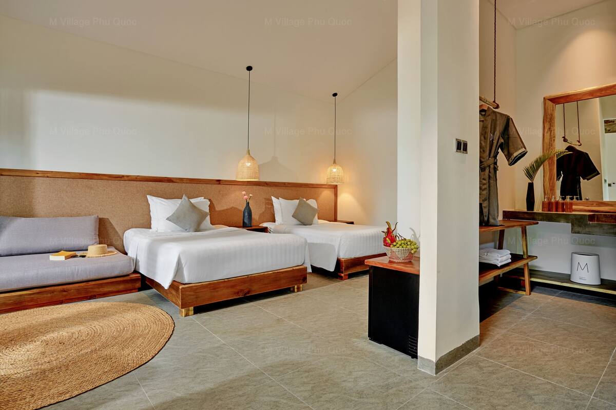 A hotel room with two beds, a sofa, a small table with a basket of fruit, and a wooden bench.