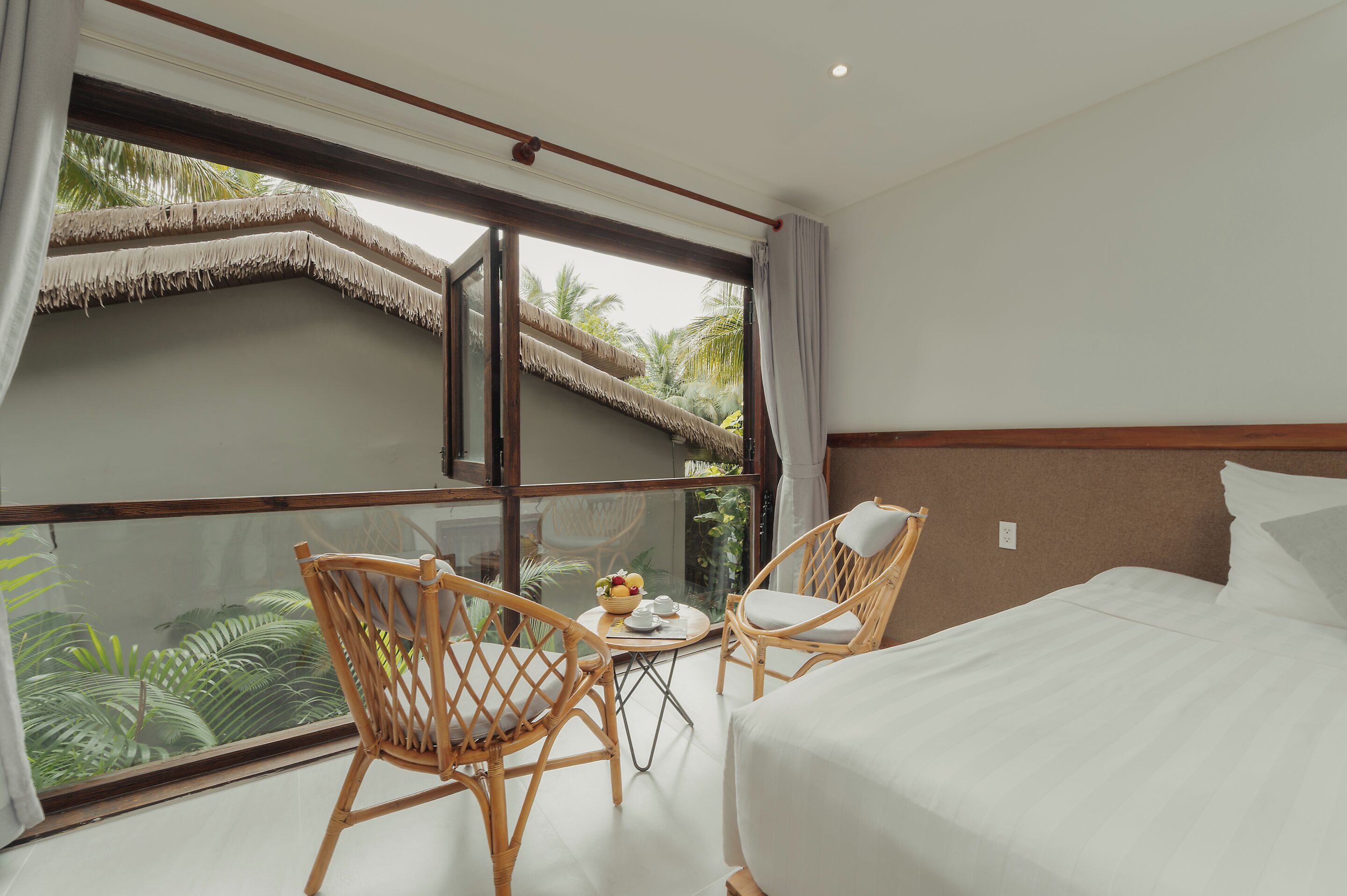 A hotel room with a large window, two wicker chairs, a small round table with a fruit bowl, and a bed with white linens.