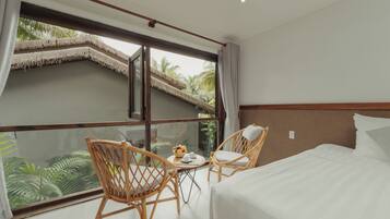 A hotel room with a large window, two wicker chairs, a small round table with a fruit bowl, and a bed with white linens.