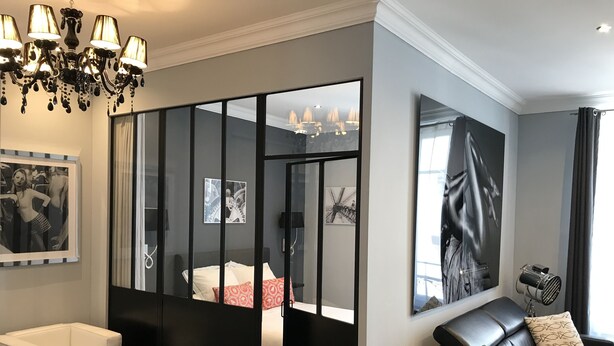 A modern living room with a black leather sofa, a white armchair, and a glass partition. There are framed artworks on the walls and a chandelier above the seating area. - CANNES APPART. LUXURY 50 m2 HYPER CENTER (Cannes)