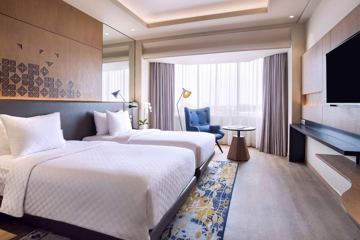 A hotel room with two beds, a TV, a desk, and a large window with curtains.