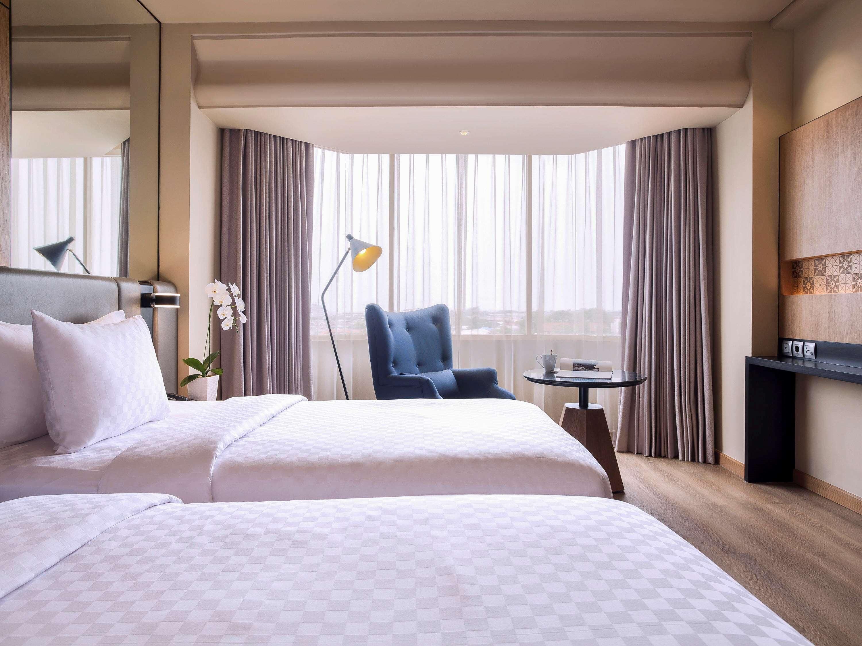 A hotel room with a large bed, a desk, a chair, a lamp, and a view through the window.