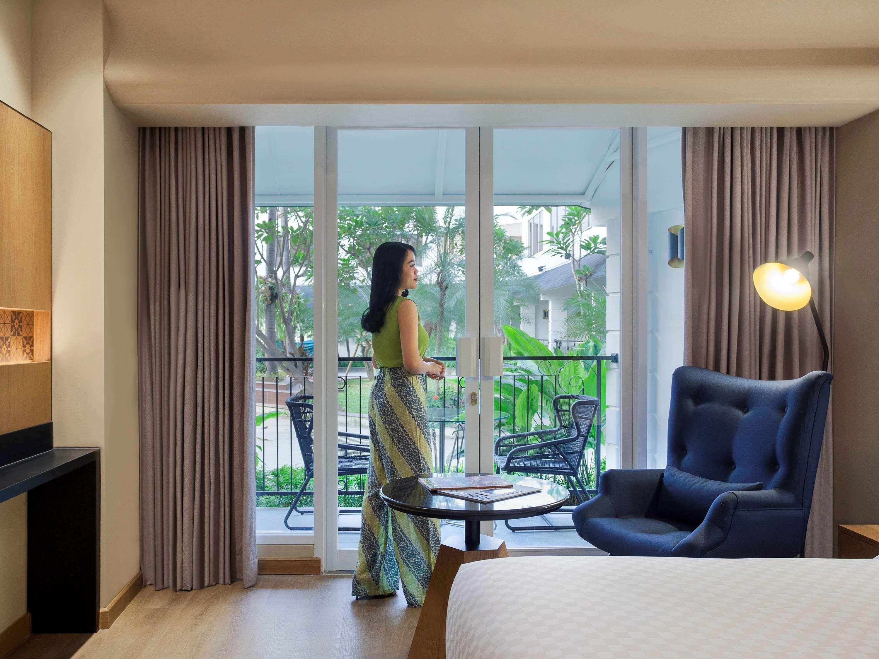 A hotel room with a bed, a chair, a table, a balcony with a view of greenery, and a large window.