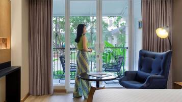 A hotel room with a bed, a chair, a table, a balcony with a view of greenery, and a large window.