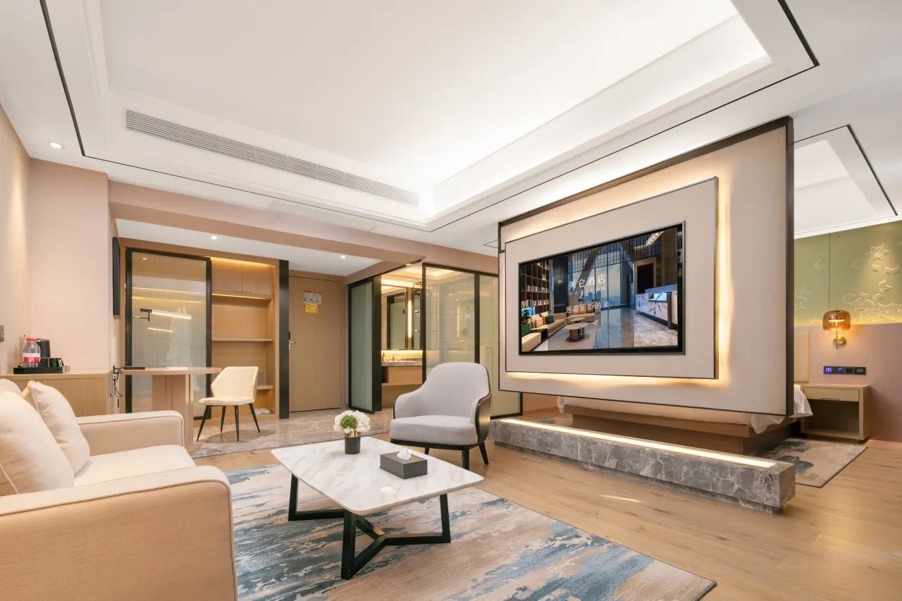 Un moderno vestíbulo de hotel con un televisor de pantalla plana montado en la pared, una zona de estar con sofás y sillones, y una mesa de centro con un florero.