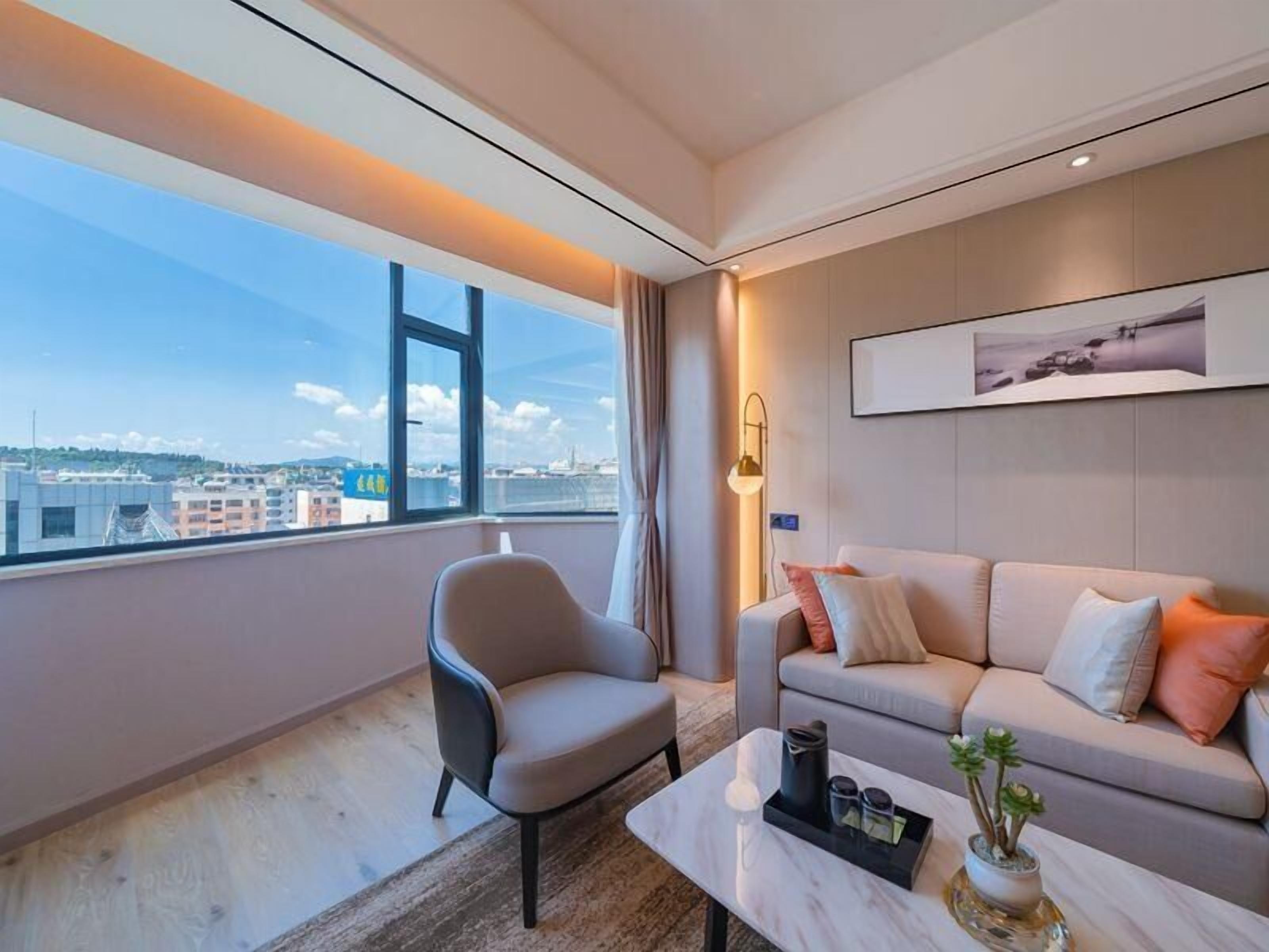 Habitació d'hotel moderna amb una finestra gran que ofereix vistes a la ciutat, un sofà confortable, un estilós butaca i una petita taula de café amb objectes decoratius.