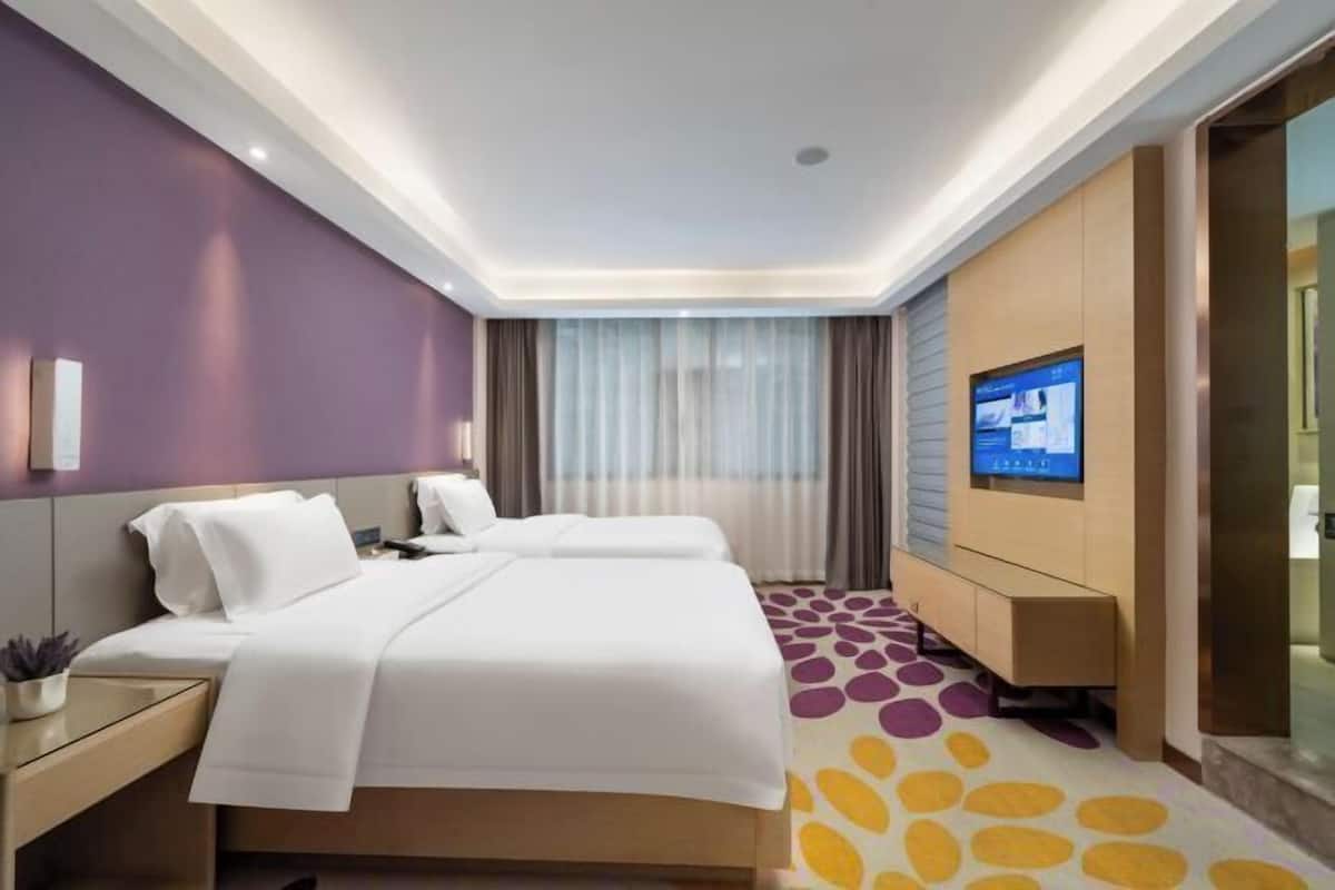 A modern hotel room with a large bed, a TV mounted on the wall, and a small desk with a lamp.