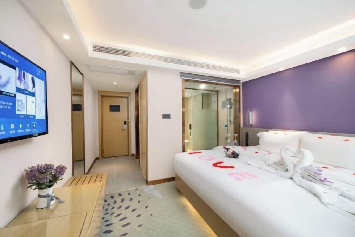 A hotel room with a bed, a TV, a small table with a vase, and a bathroom visible through a glass door.