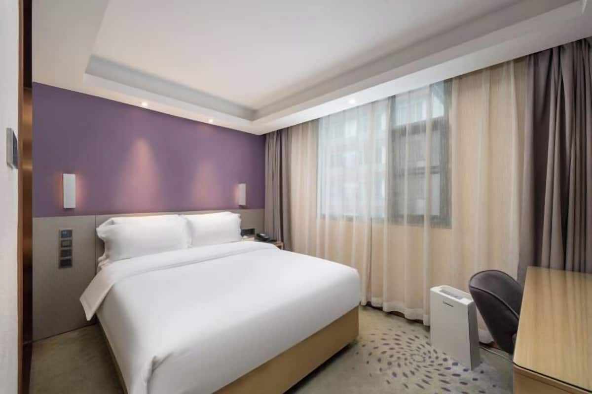 A hotel room with a bed, a desk, a chair, a nightstand, and a window with curtains.