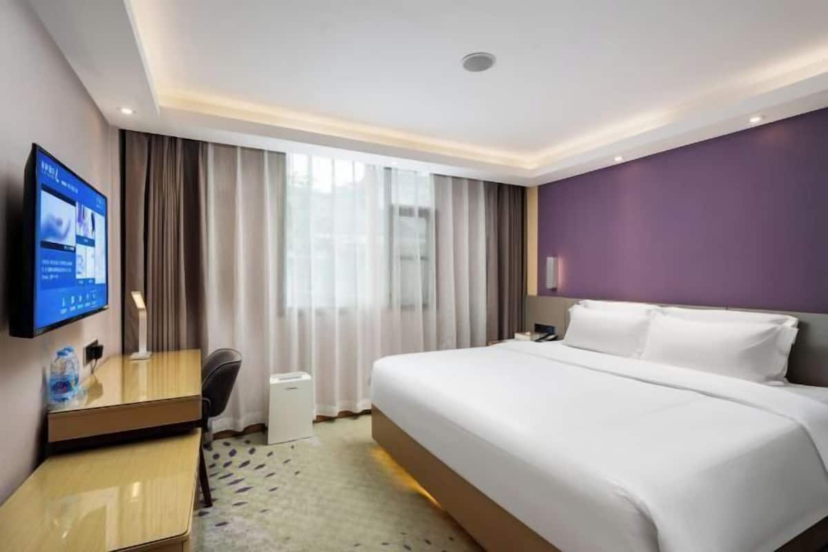 A hotel room with a large bed, a desk with a computer, a TV, and a window with curtains.