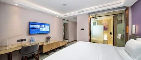 A hotel room with a bed, desk, chair, television, and a bathroom visible through a glass door.