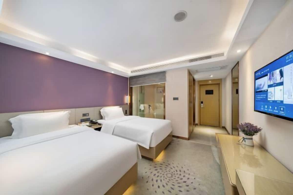A hotel room with two beds, a desk, a flat-screen TV, and a bathroom visible through a glass partition.