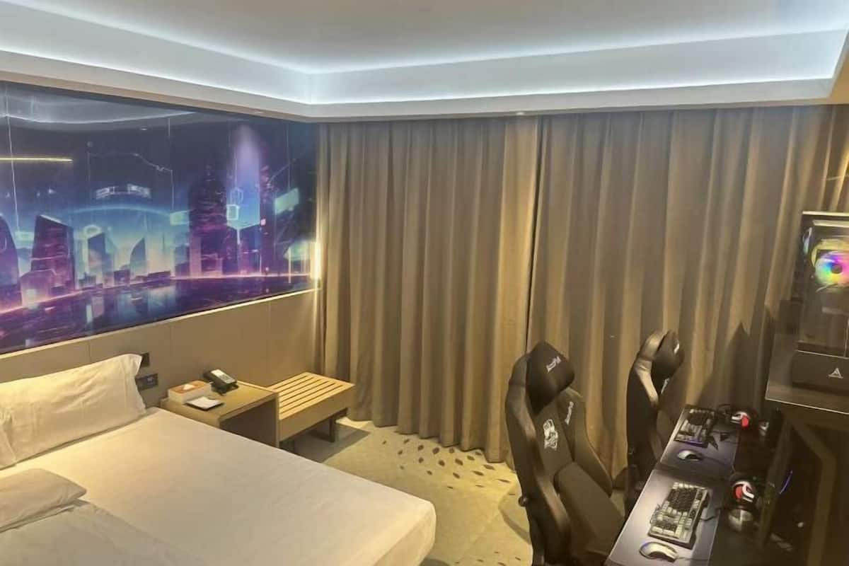 A hotel room with a bed, a desk with a computer, a gaming chair, a TV with a cityscape on the wall, and a curtain.