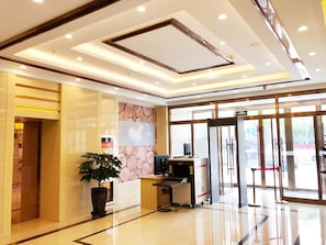 Eine moderne Hotellobby mit einer Rezeption, einer großen Topfpflanze und einem Glastür-Eingang.