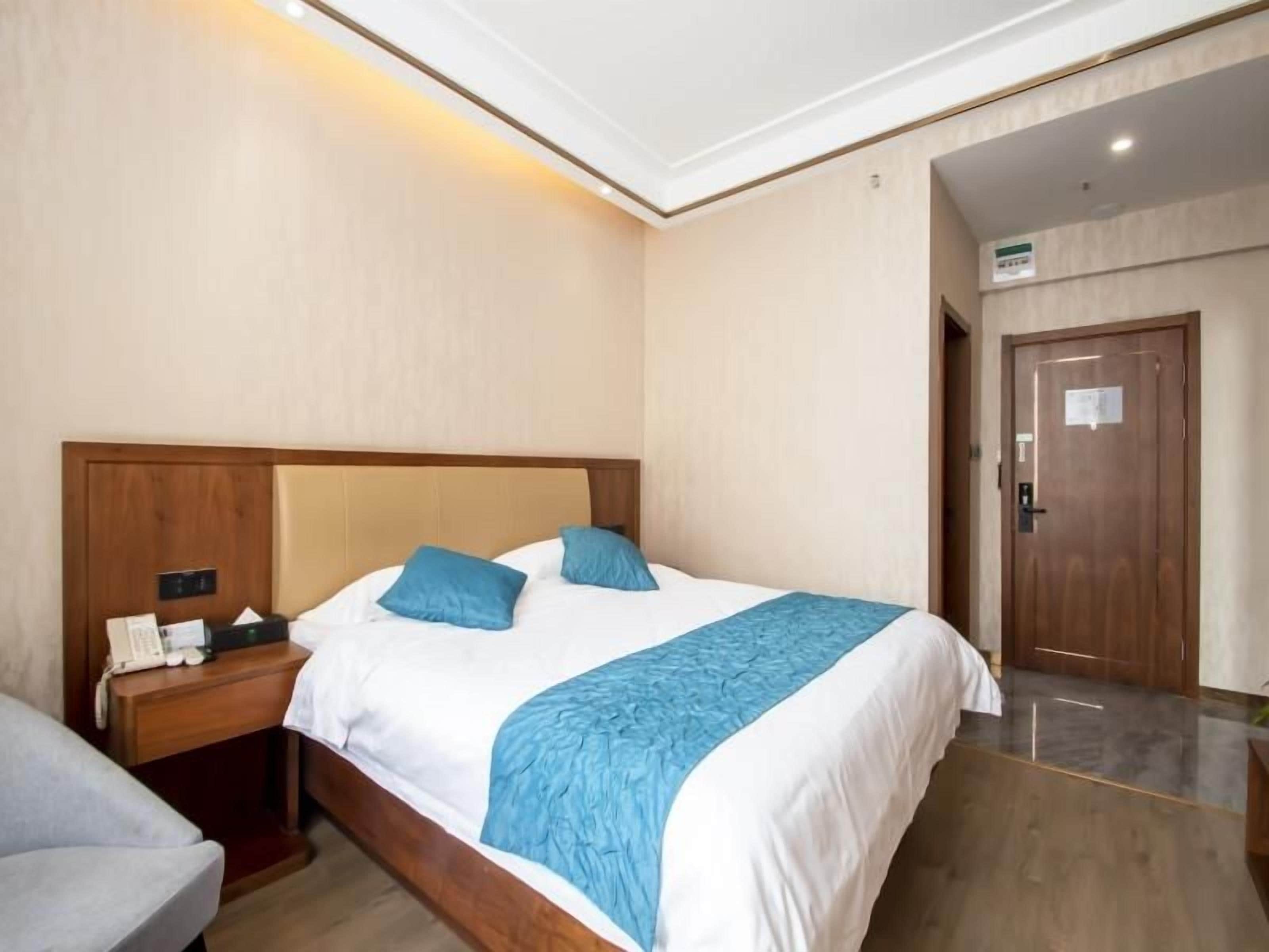 Et hotelværelse med sengegavl i træ, seng med hvide sengetøj og blå detaljer, et natbord med telefon og papirer, samt en dør med et skilt.